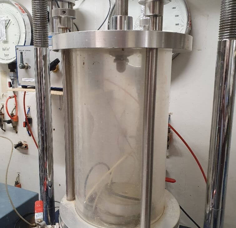 A laboratory triaxial test apparatus with a transparent cylindrical chamber, metal supports, pressure gauges, and connected red tubes, used for testing soil samples under controlled pressure conditions.