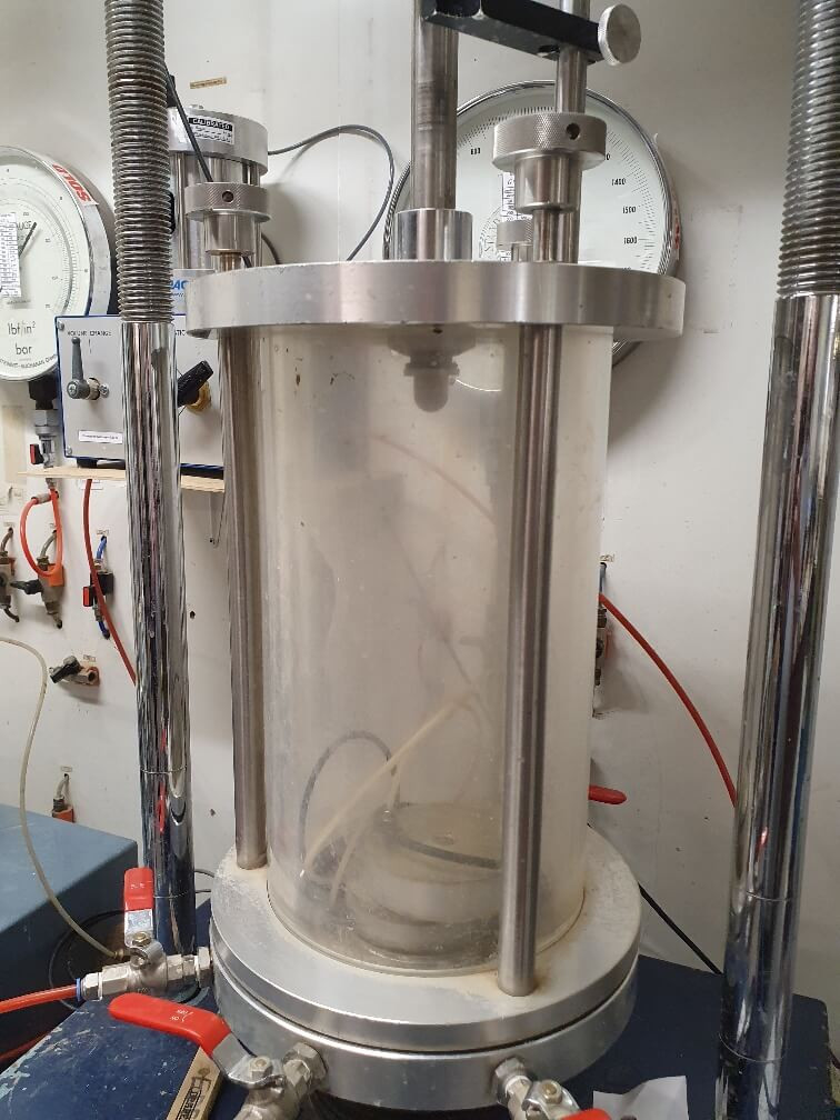 A laboratory triaxial test apparatus with a transparent cylindrical chamber, metal supports, pressure gauges, and connected red tubes, used for testing soil samples under controlled pressure conditions.