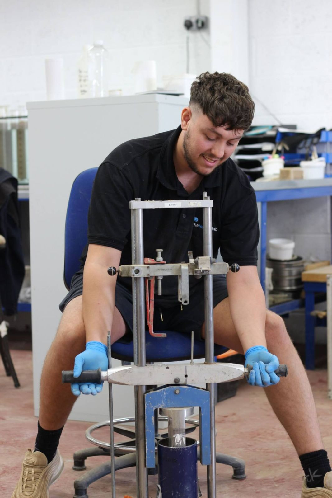 A man wearing blue gloves and a black shirt operates a manual laboratory press in a workshop, sitting on a chair and handling the equipment with both hands. Laboratory tools and shelves are visible in the background.