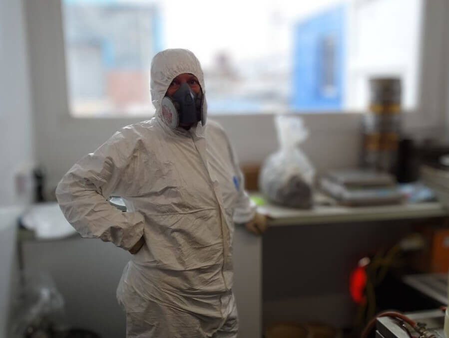 A person wearing a white hazmat suit, gloves, and a respirator stands in a laboratory, with equipment and supplies visible on benches in the background.
