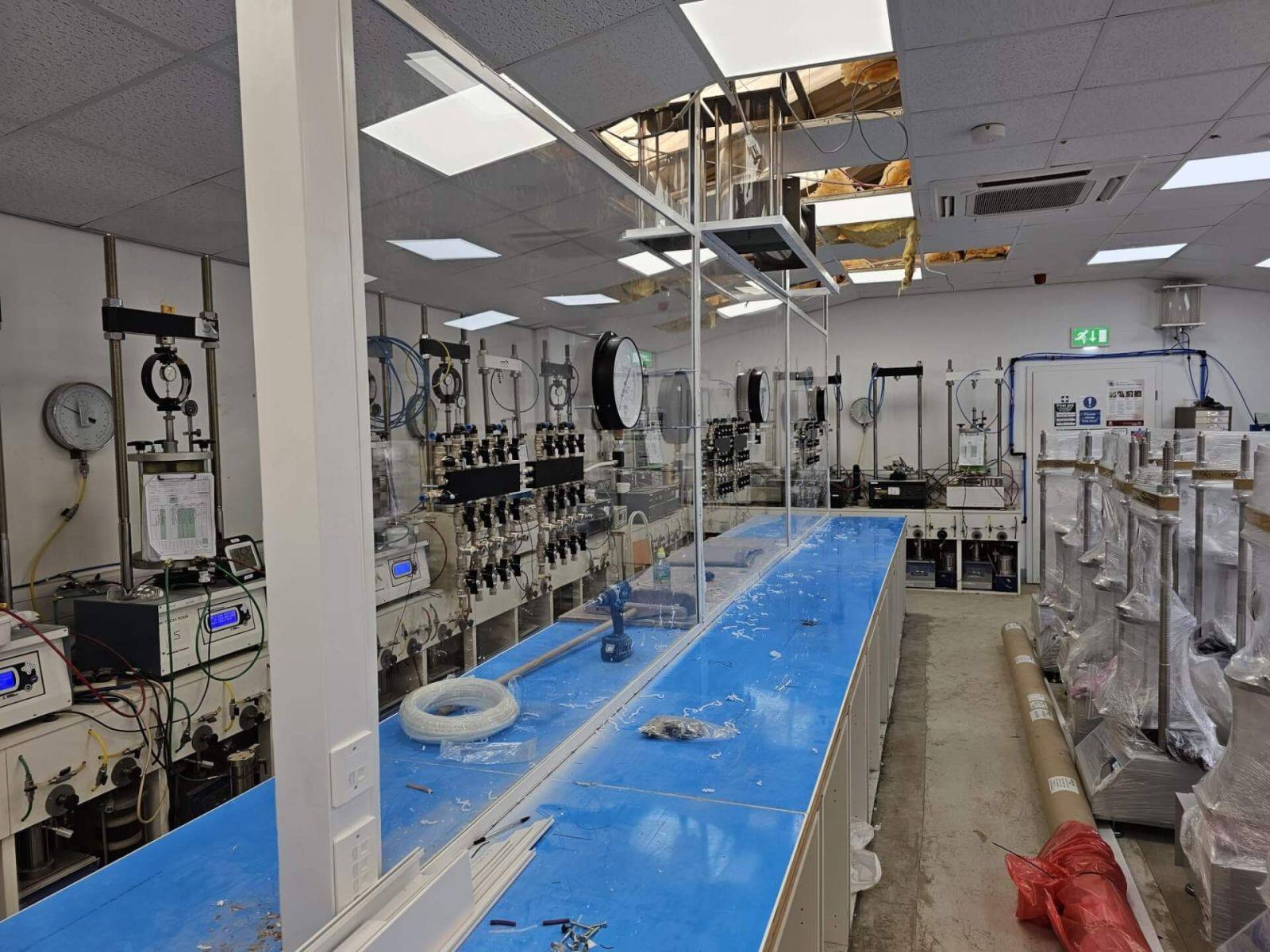 A laboratory with blue workbenches, scientific equipment, gauges on the walls, and a partially damaged ceiling exposing insulation. The room is well-lit with overhead lights and has various supplies and boxes along the right side.