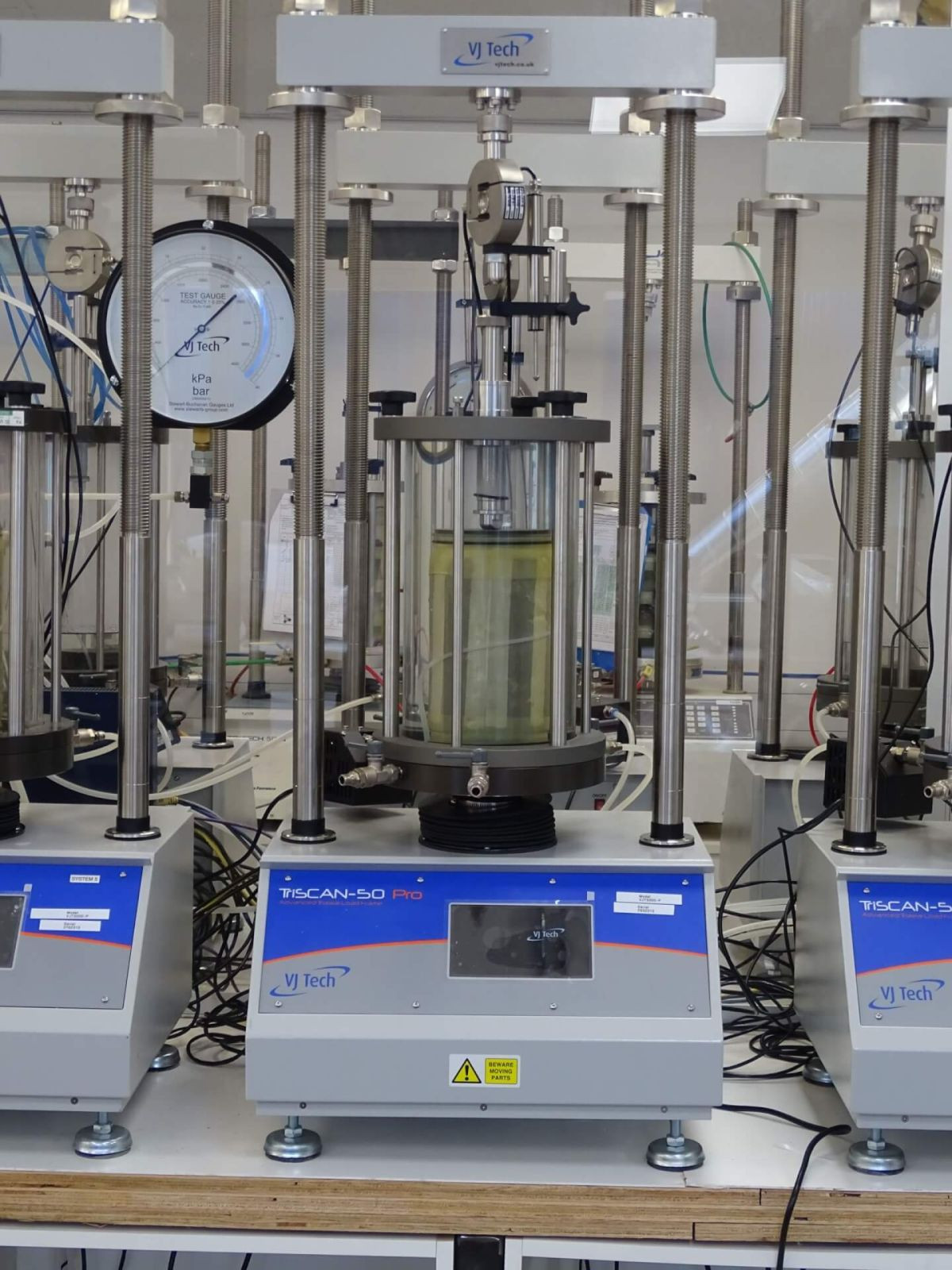A laboratory triaxial testing machine with a transparent cylindrical cell, pressure gauges, and digital controls, used for soil mechanics experiments; wires and equipment are visible around it.