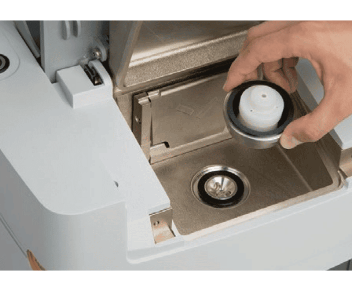 A hand removes a round water tank filter from a compartment in a coffee machine, revealing the inside of the water reservoir.